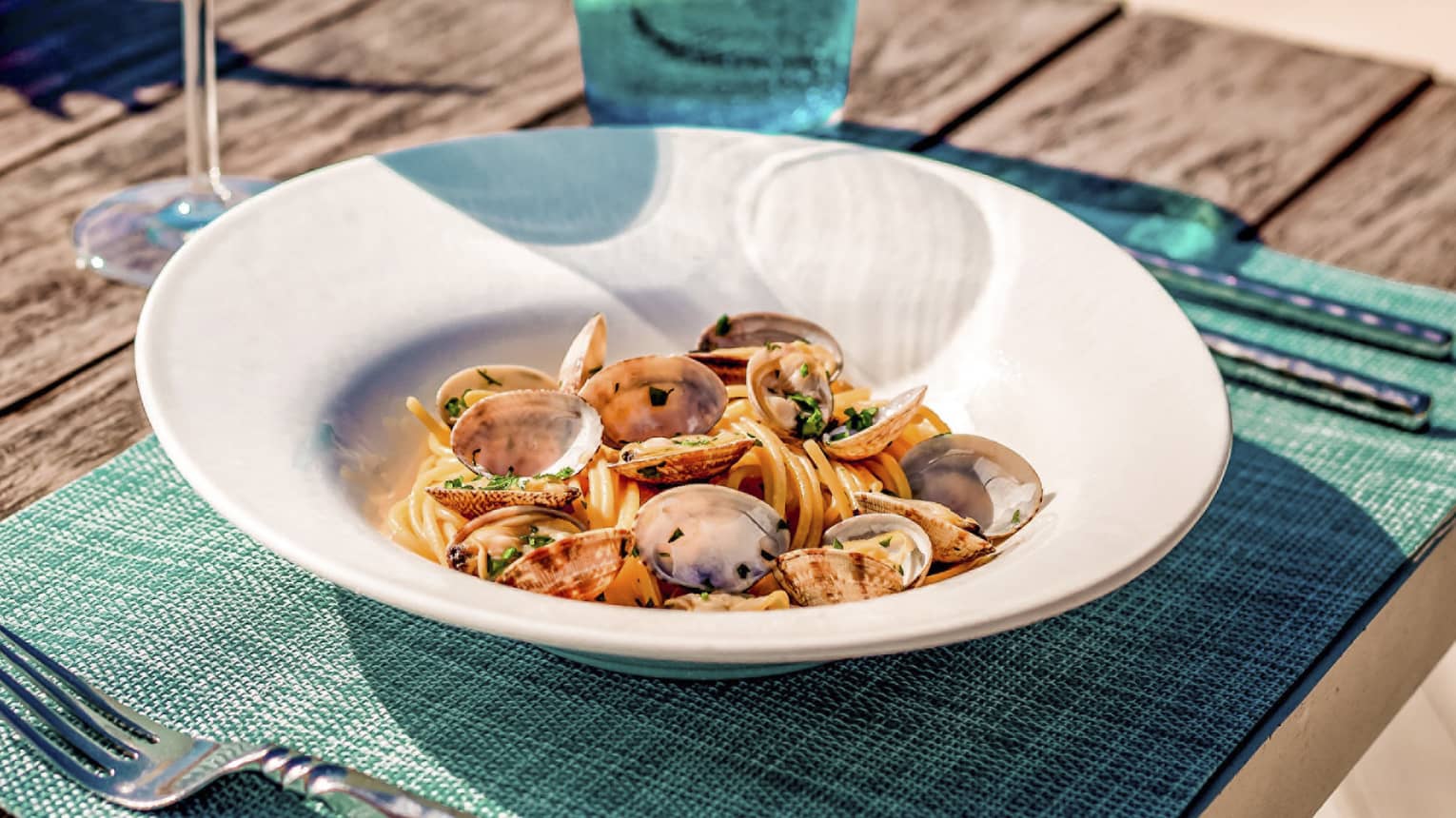 Spaghetti alle vongole on wooden picnic table with turquoise placemat and water glass, clear wine glass, conch shell