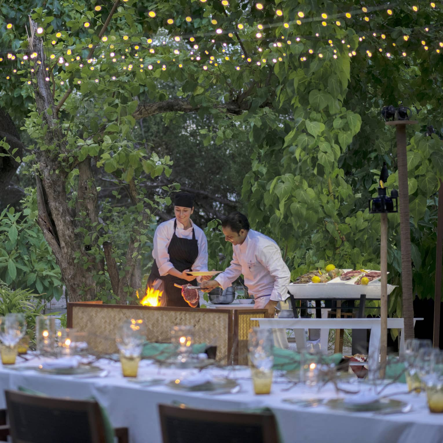 Chefs put large steak on flaming grill by long outdoor dining table, patio lights at night
