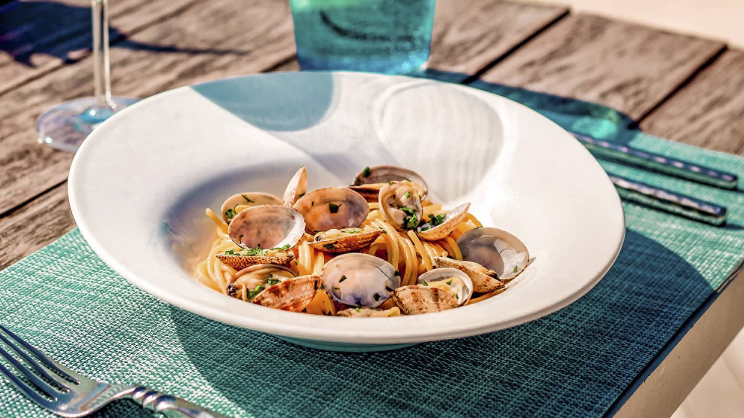 Spaghetti alle vongole on wooden picnic table with turquoise placemat and water glass, clear wine glass, conch shell
