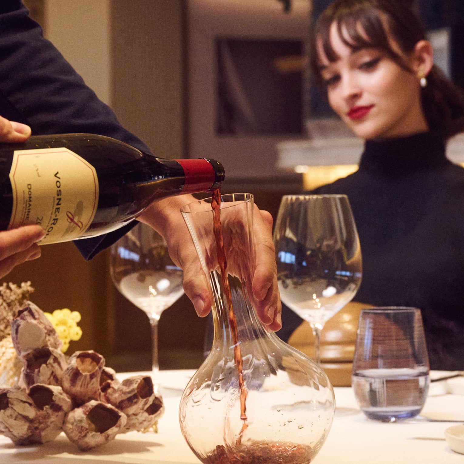 Suited server decants red wine at table with seated guest watching