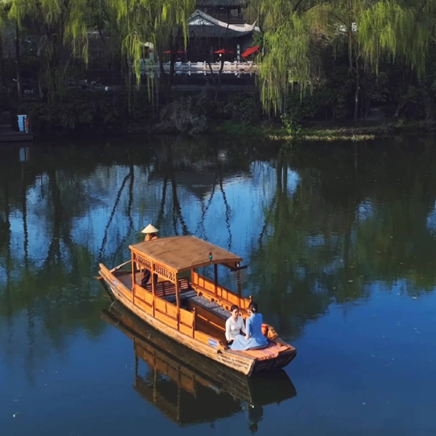 Facing each other, two guests sit in the front of a small wooden boat on a peaceful lake as a third person steers the craft.