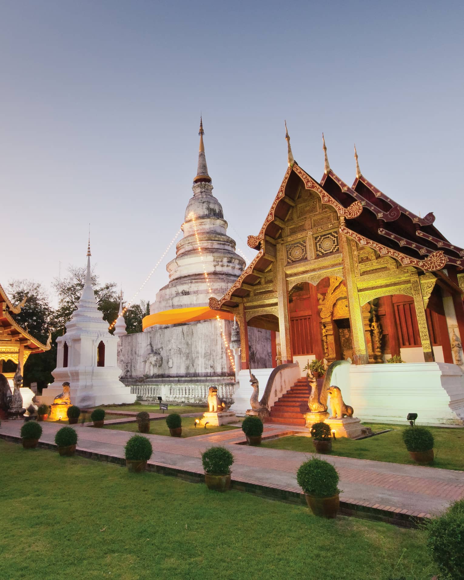 Two temples with gabled roofs and intricate carvings uplit in gold, between them a white domed building and stone monument.