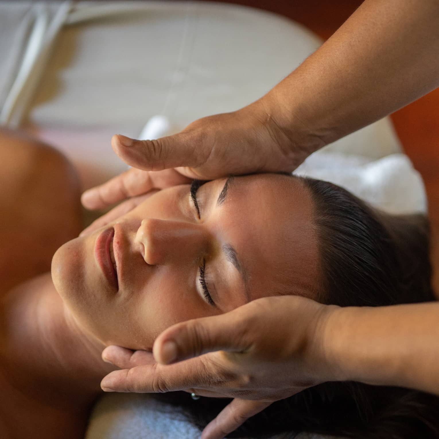 Woman lays on massage table with eyes closed as hands massage her cheeks and chin