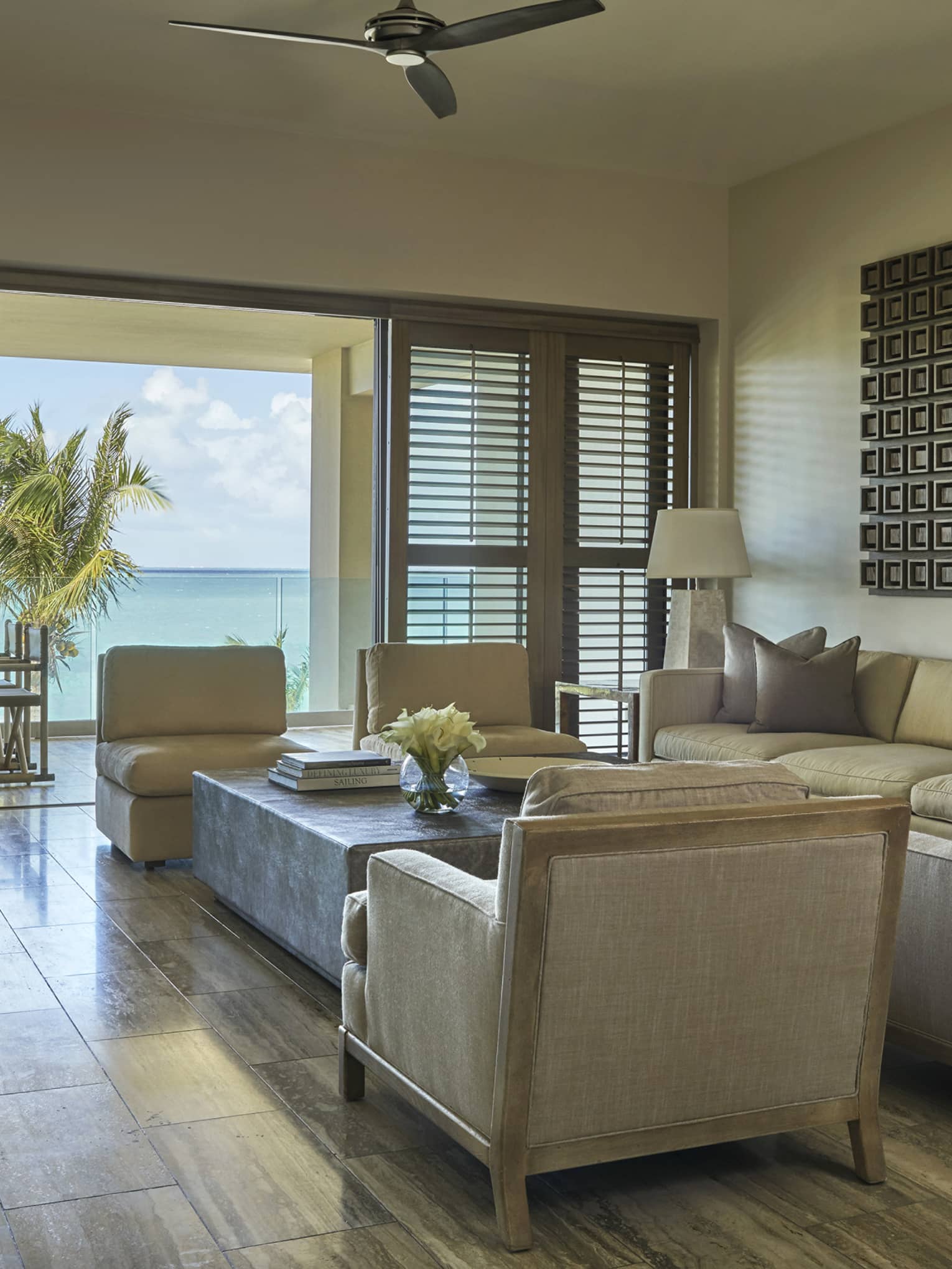 Bright living room with beige armchairs, sofa and ocean-view terrace framed by palm trees