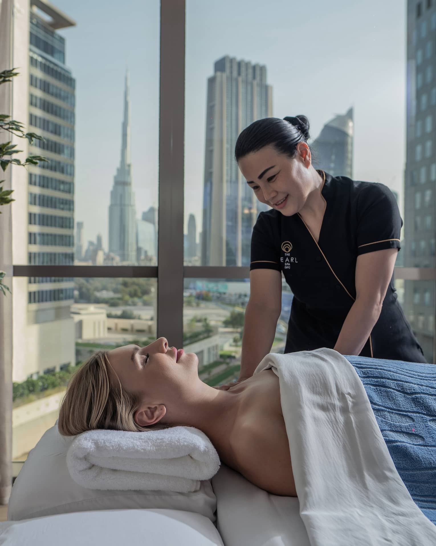 A guest enjoys a massage from a Hotel employee. Behind them, a window frames tall glass skyscrapers and clusters of trees.