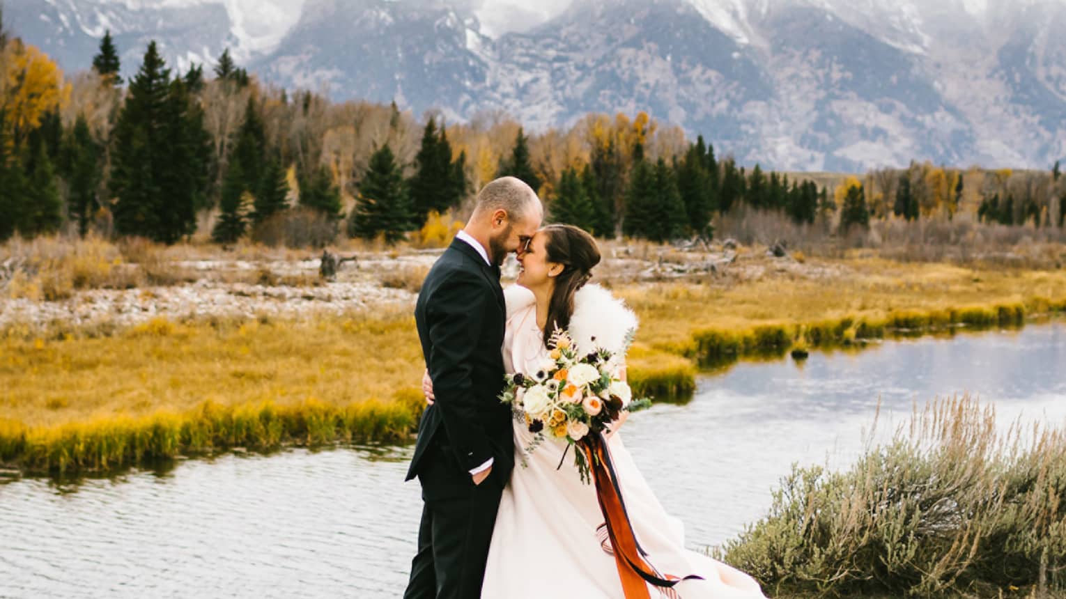 Man in suit and woman in wedding dress with bouquet embrace outdoors with backdrop of river and mountains