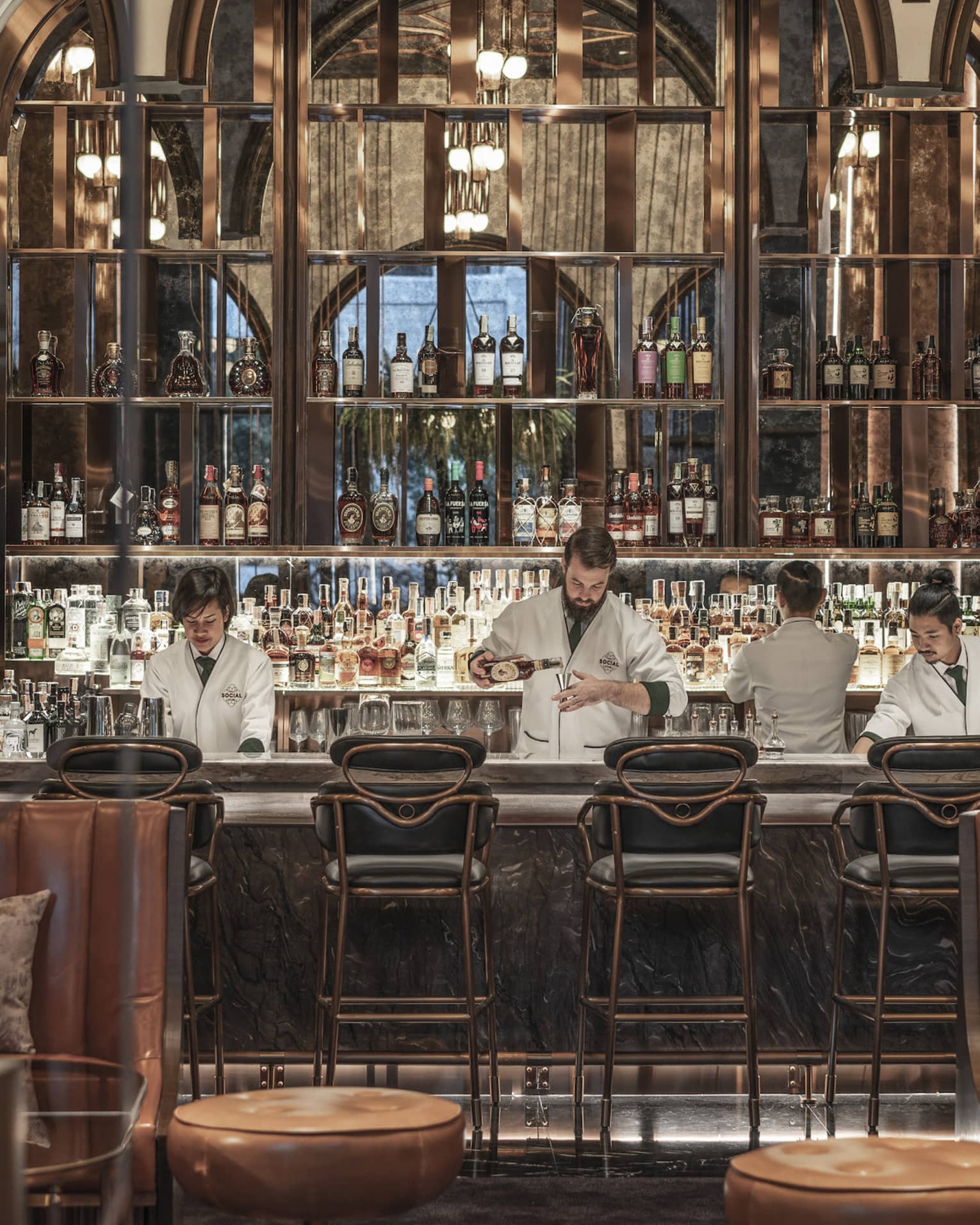 Five bartenders in white shirts prep behind a bar, brown leather booth seating in foreground