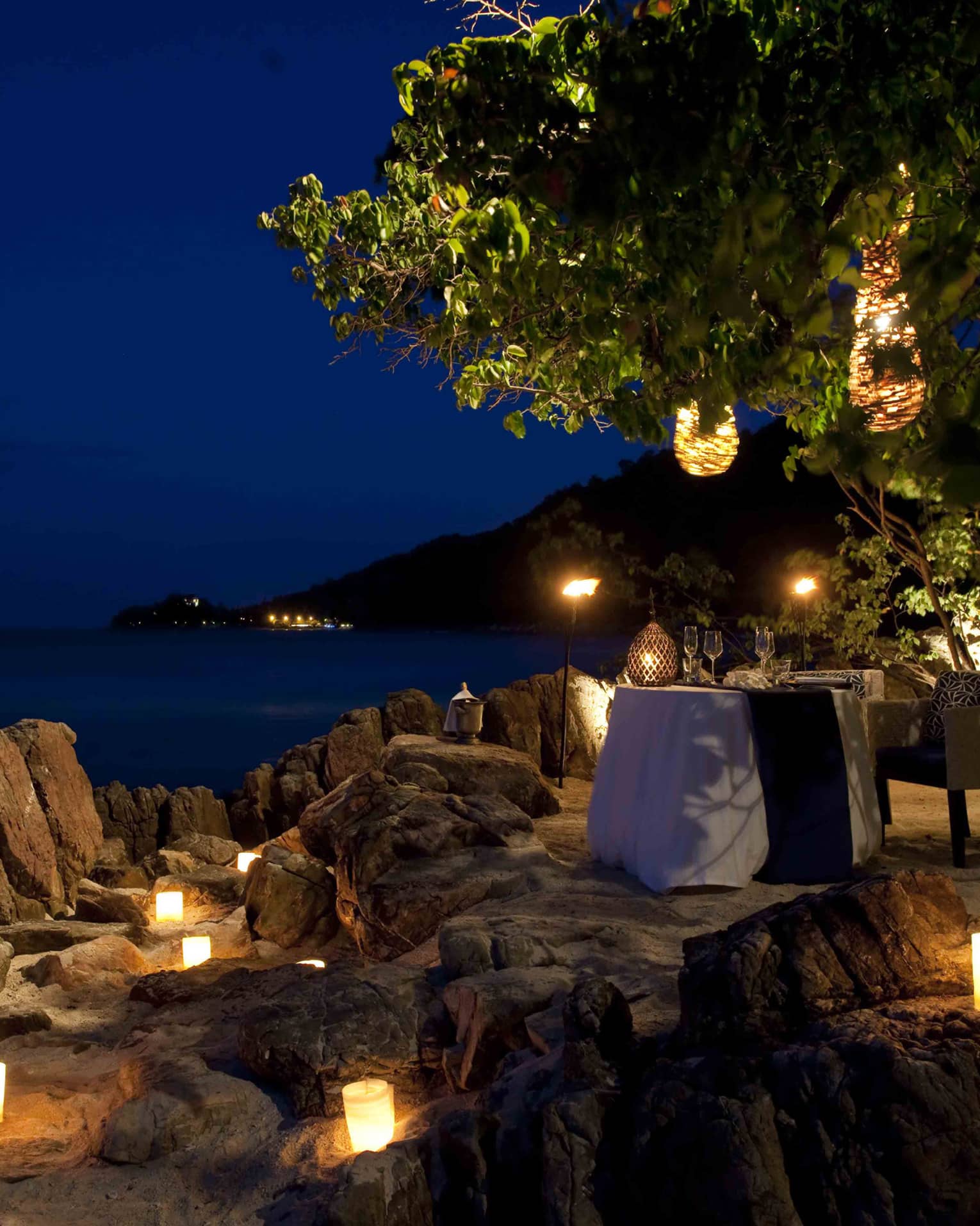 Candles glow on rocks near private dining table on beach at night