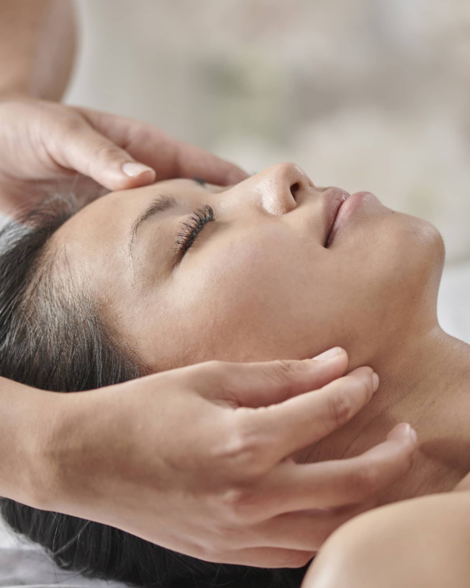 Masseuse massages woman's face as she lies face up on table under white sheet