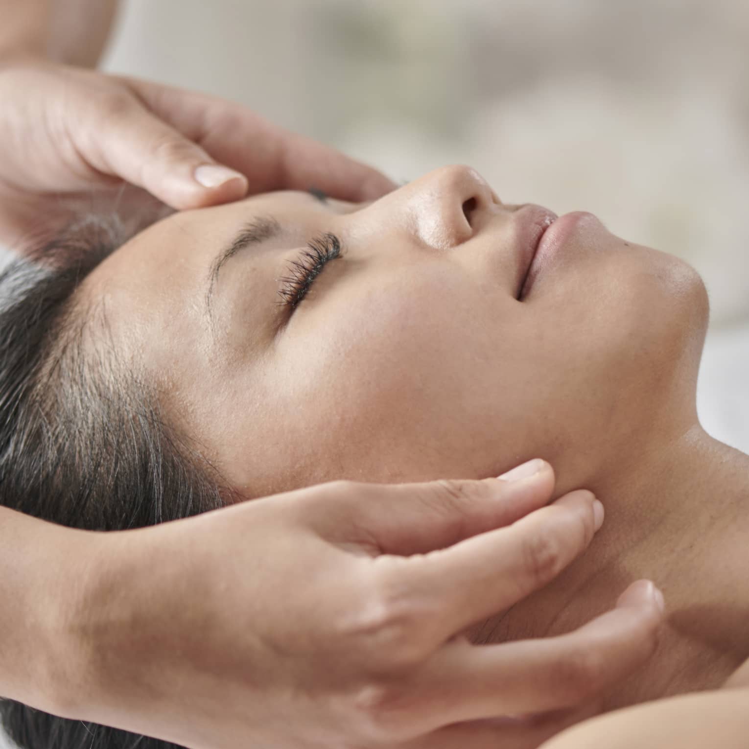 Masseuse massages woman's face as she lies face up on table under white sheet