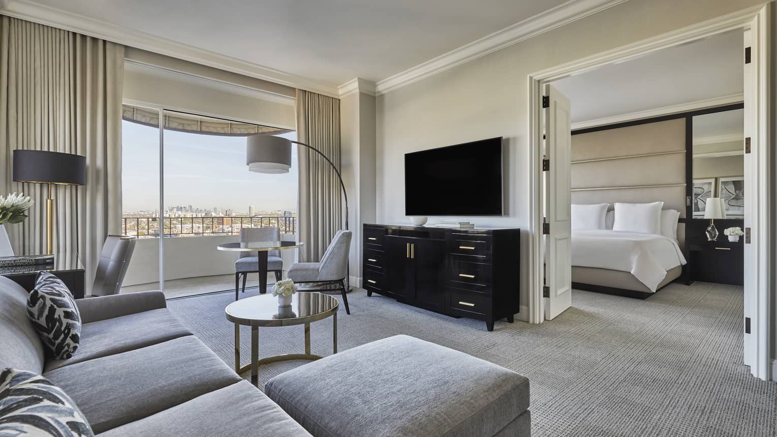 A hotel room with a grey couch, dresser, tv, lamp, small table, bed with white sheets, and large window.