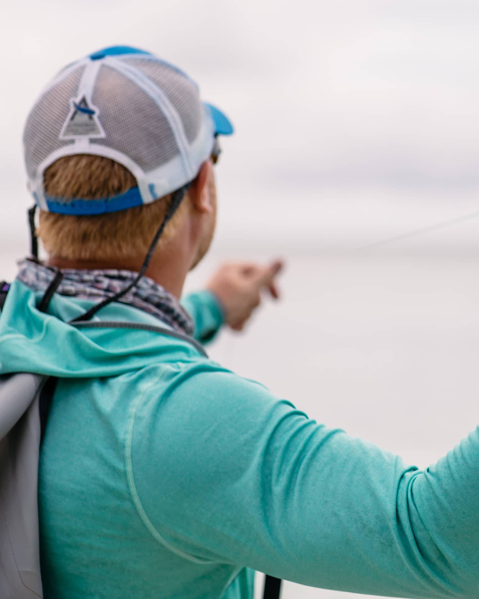 A fisherman facing away holds his fishing line in his left hand and draws back his rod with his right, readying to cast. 