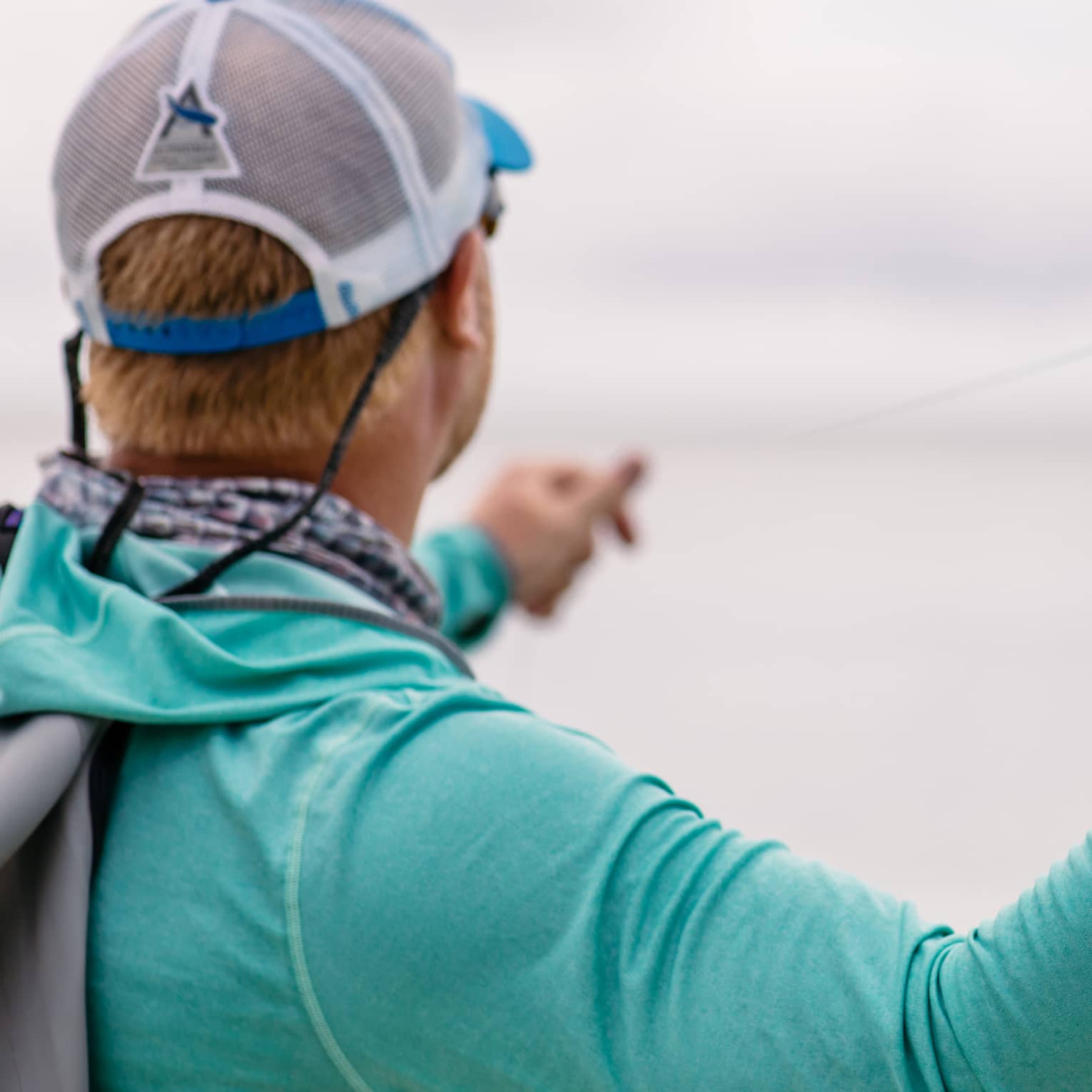 A fisherman facing away holds his fishing line in his left hand and draws back his rod with his right, readying to cast. 