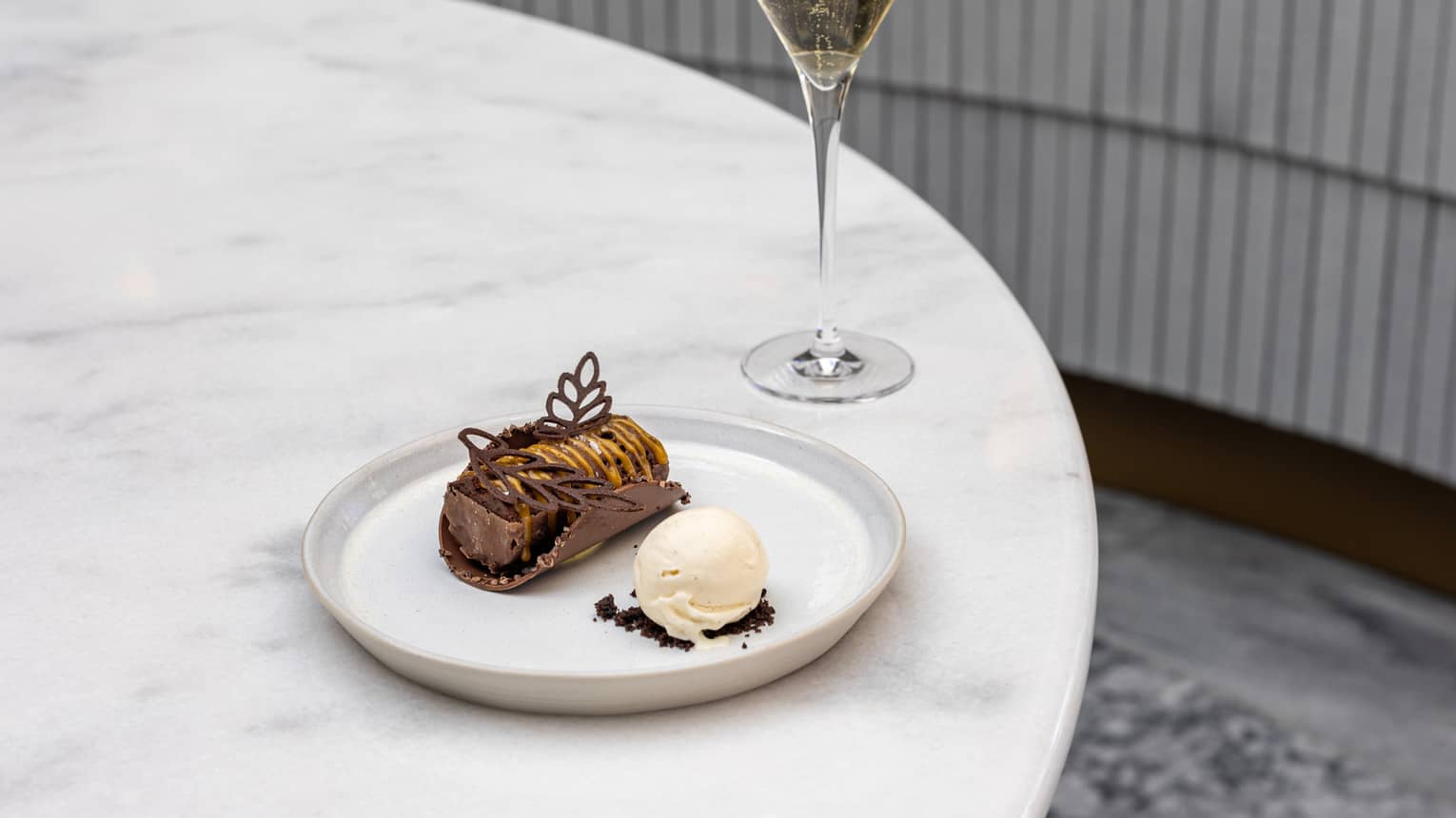 An oblong golden-brown dessert with leaf-shaped decorations on top on a white plate along with a scoop of ice cream sits on a white table next to a pale drink in a clear stemmed glass
