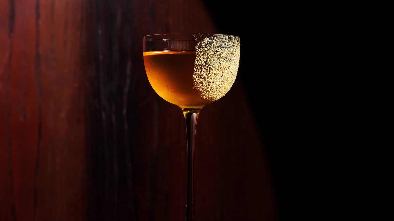 A tall stemmed red wine glass holds a golden-colored cocktail with gold dust sprinkled on the right side of the glass