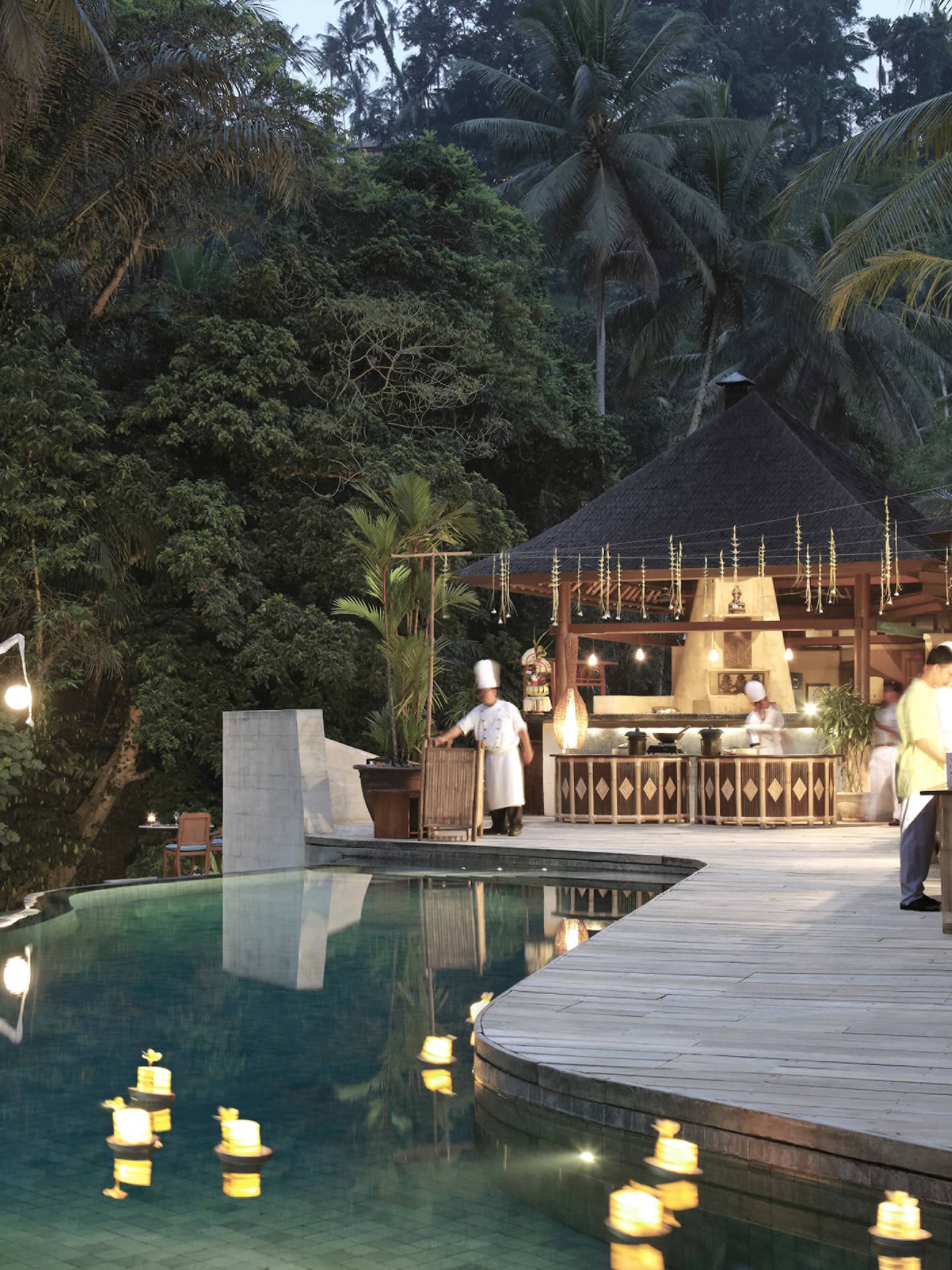 An infinity pool looking out towards the dense jungle; chefs bustle around the outdoor kitchen on the poolside deck.