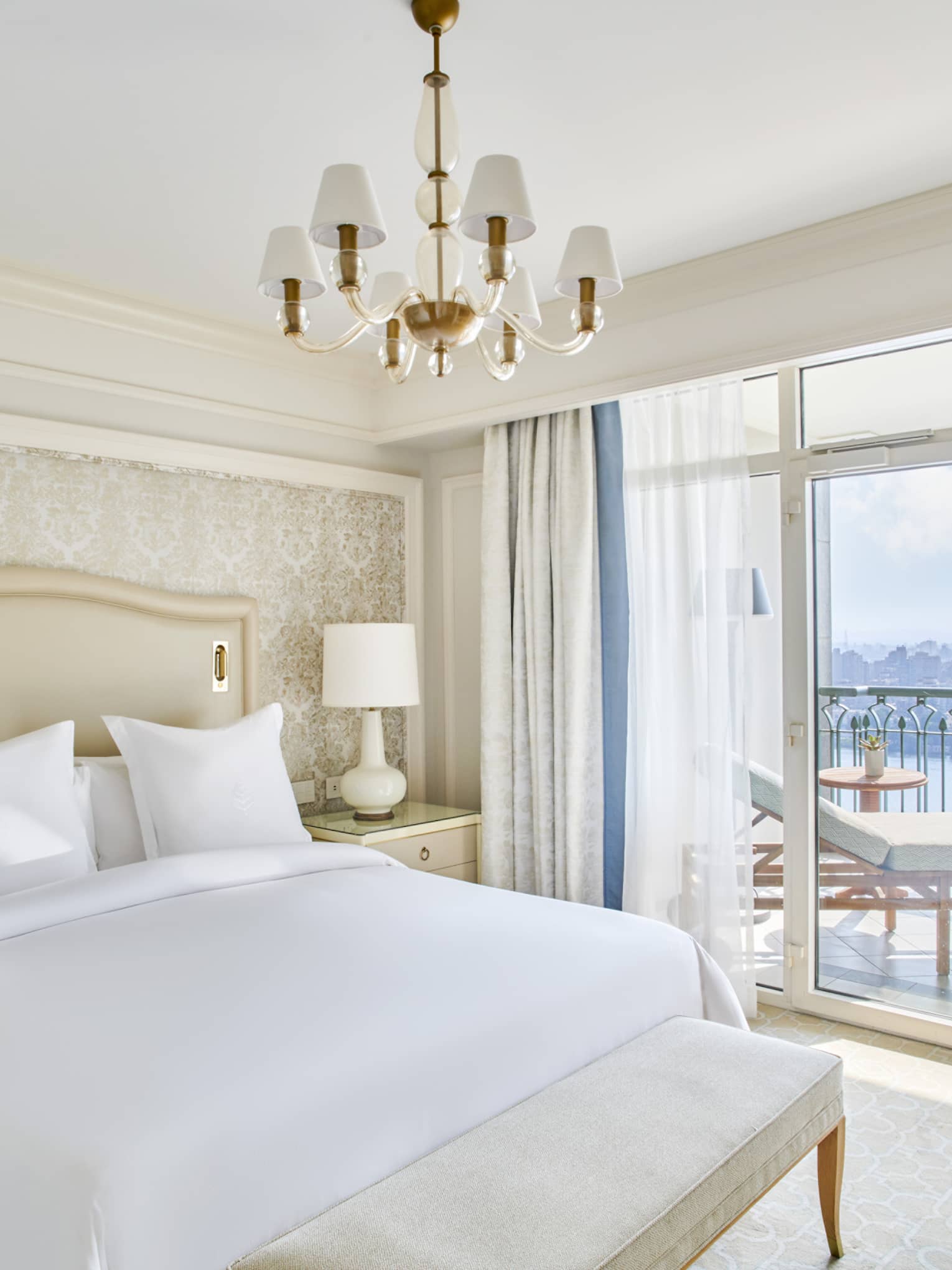 Guest bedroom with king bed, white linens and cream tufted headboard; foot bench, sliding glass door to balcony with Nile River views