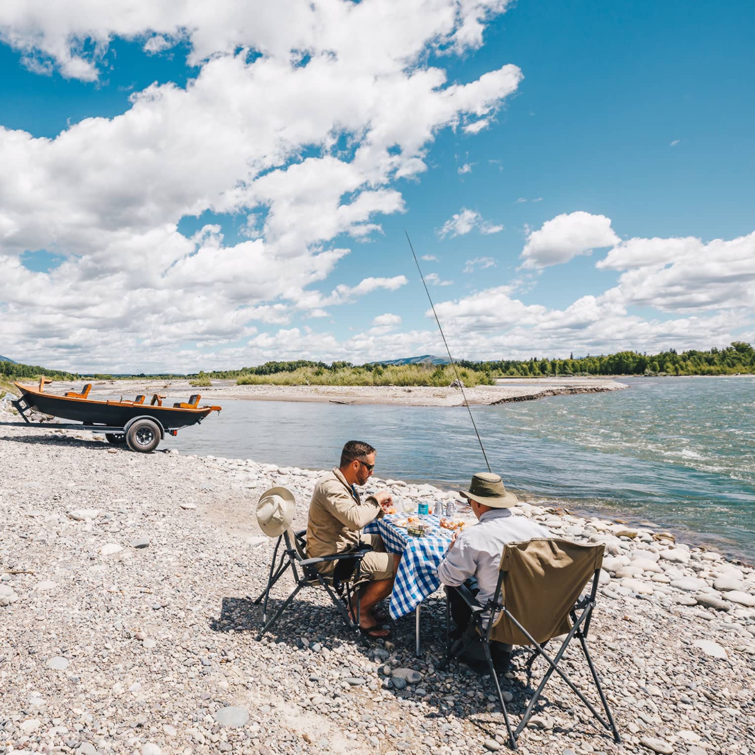 Two fishermen sit at a small table for a lakeside picnic, their boat hitched to their SUV in the background