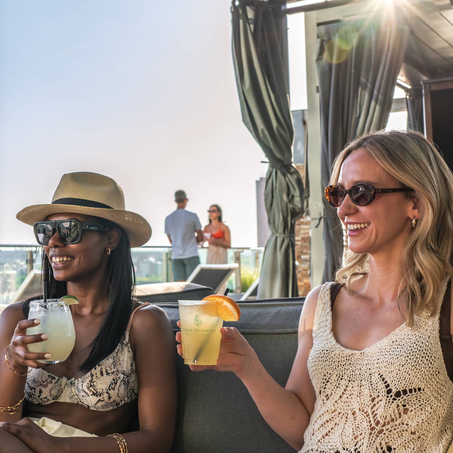 Two smiling friends holding iced beverages lounge on a cabana sofa, a bright burst of sunlight streaming through behind them.