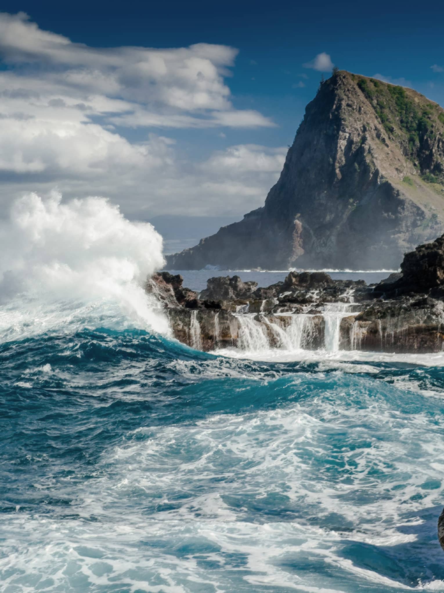 Crashing waves strike the shore of a volcanic island. A mountain looms in the background against a partially-cloudy sky.