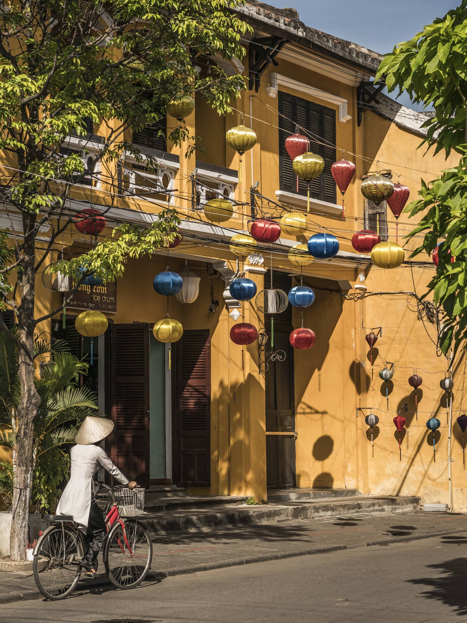 A woman biking down a historic street in Hoi, Viet Nam with lanterns along the side of a house 