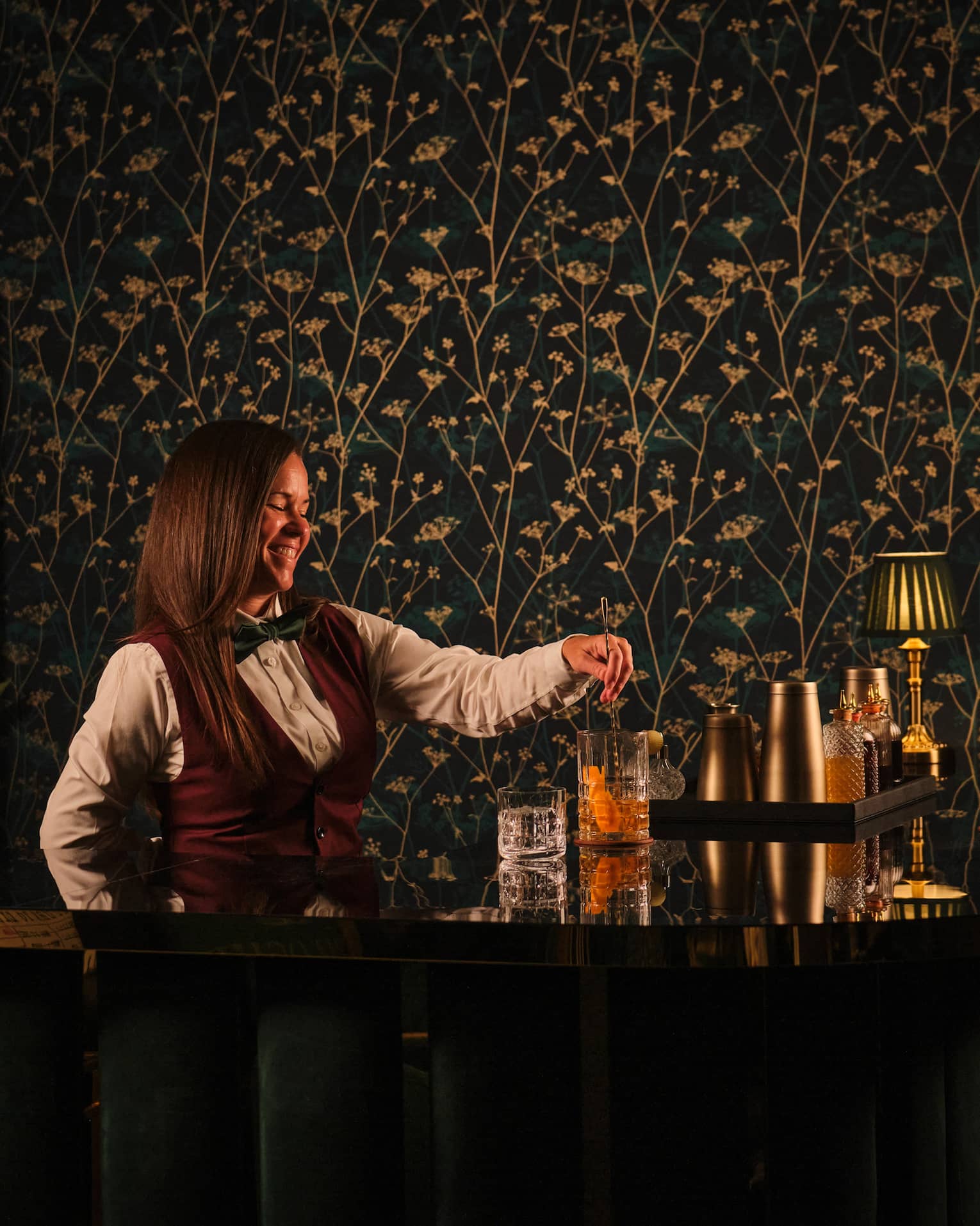 Bartender wearing white shirt, burgundy vest and black bowtie pours a cocktail from behind the bar