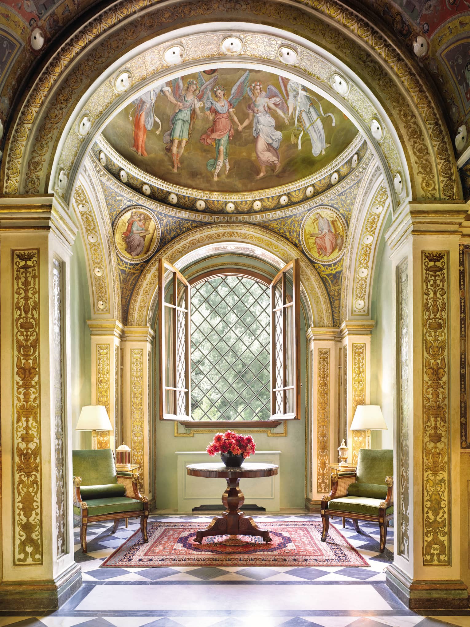 Green velvet armchairs around wood table with flowers, open window, gold ornate pillars