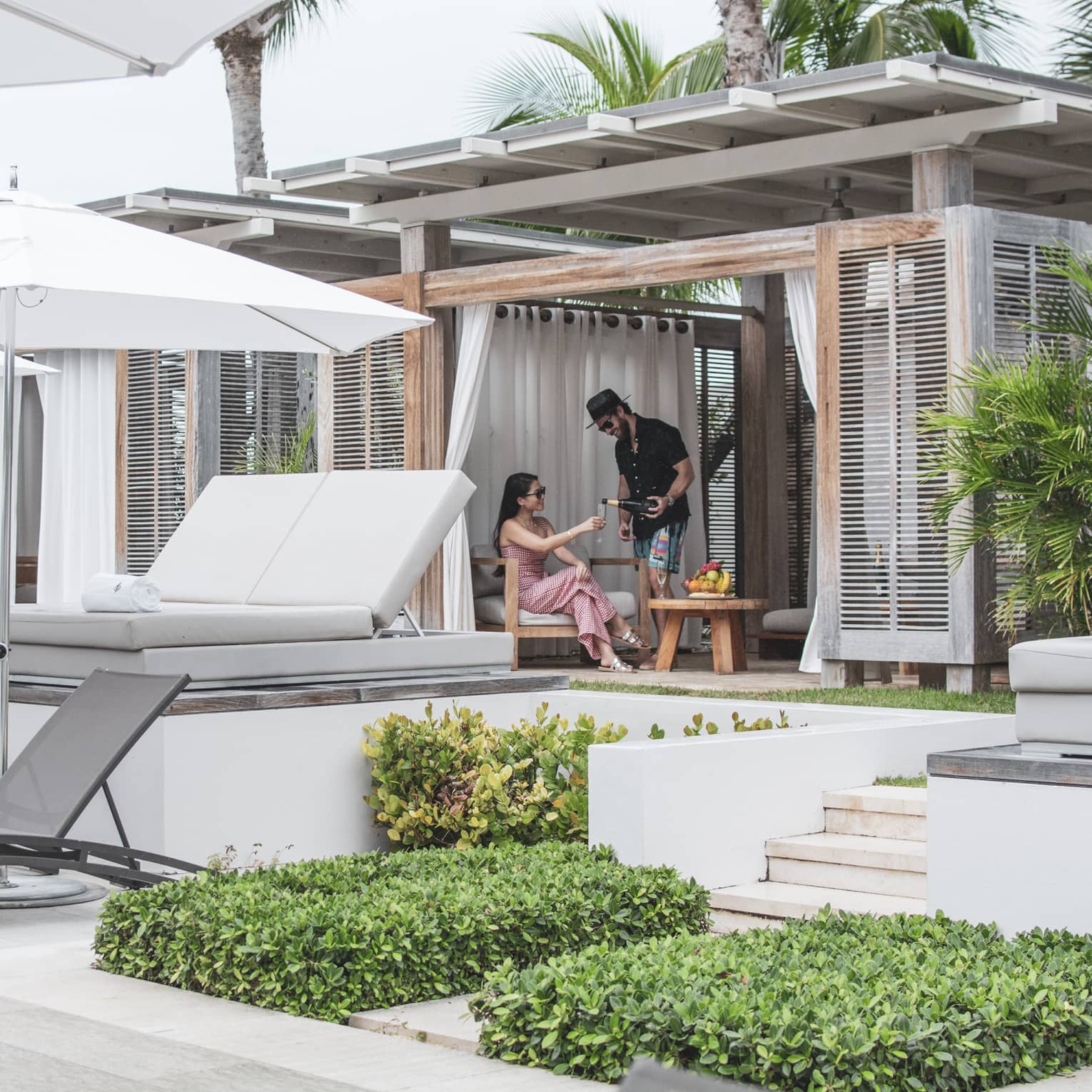 Two talk to each other in a covered pool cabana with a row of white daybeds and a row of grey lounge chairs and white umbrellas in front of them