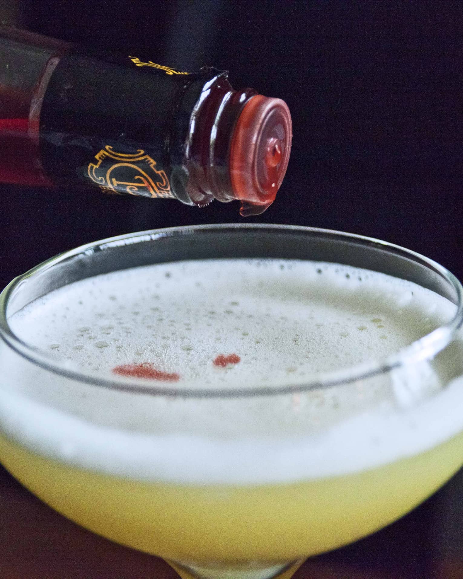 Close-up of martini glass with yellow cocktail, white foam, bottle with red liqueur dripping out