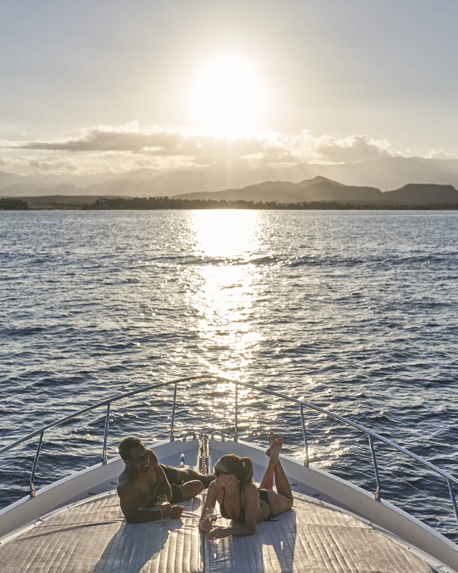 Couple wearing swimsuits relaxes on front yacht deck as sun sets over ocean