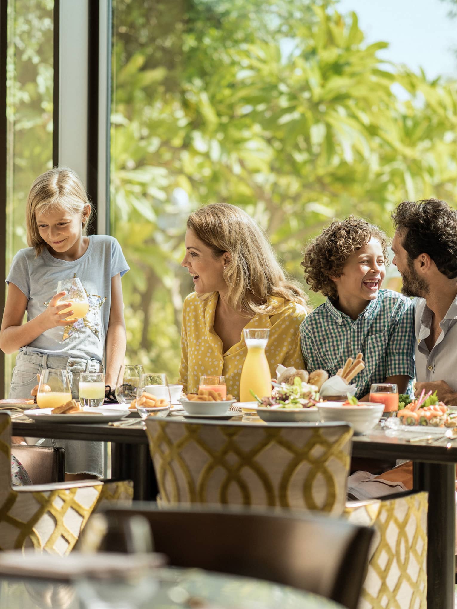 A family of five eating breakfast on the same side of the table, with trees in the background.