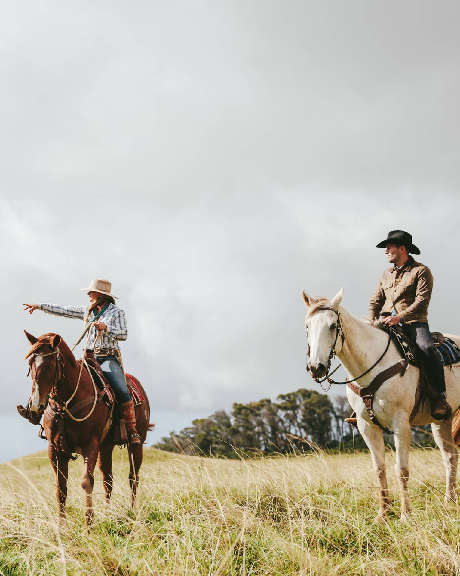 A couple riding horses, looking out at the long grass fields, with grassy hills and trees in the distance on a cloudy day.