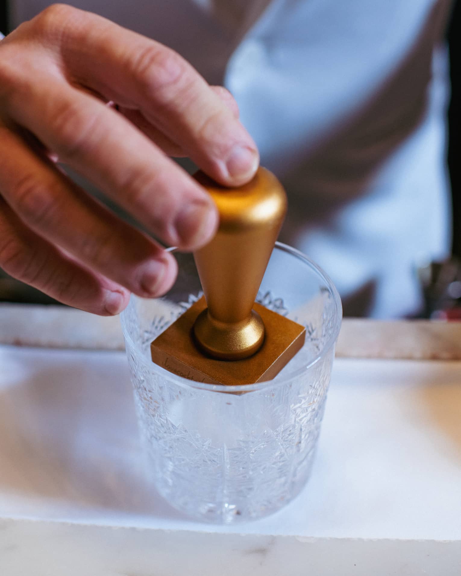 Close-up of a hand embossing an ice cube using a brass ice stamp in a clear cut-crystal lowball cocktail tumbler.
