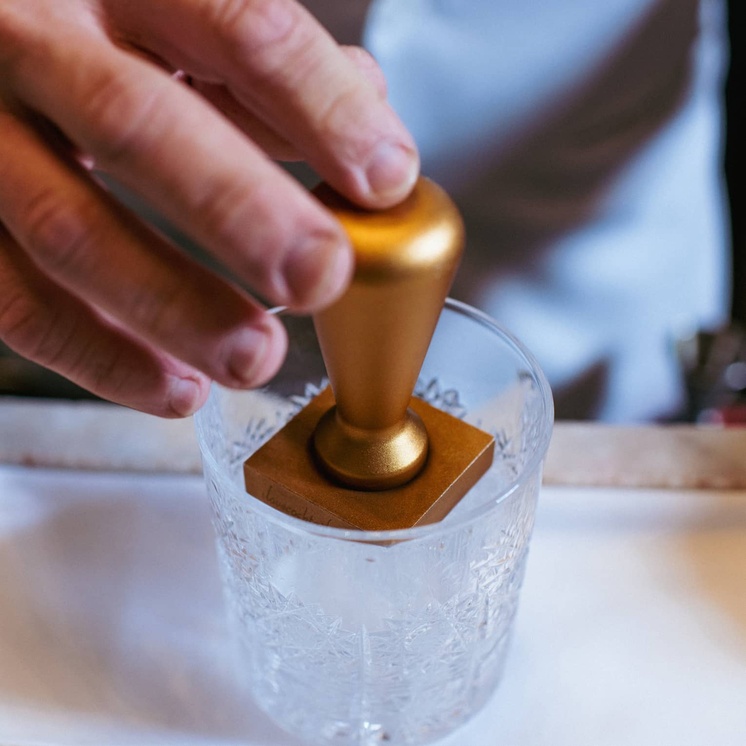 Close-up of a hand embossing an ice cube using a brass ice stamp in a clear cut-crystal lowball cocktail tumbler.