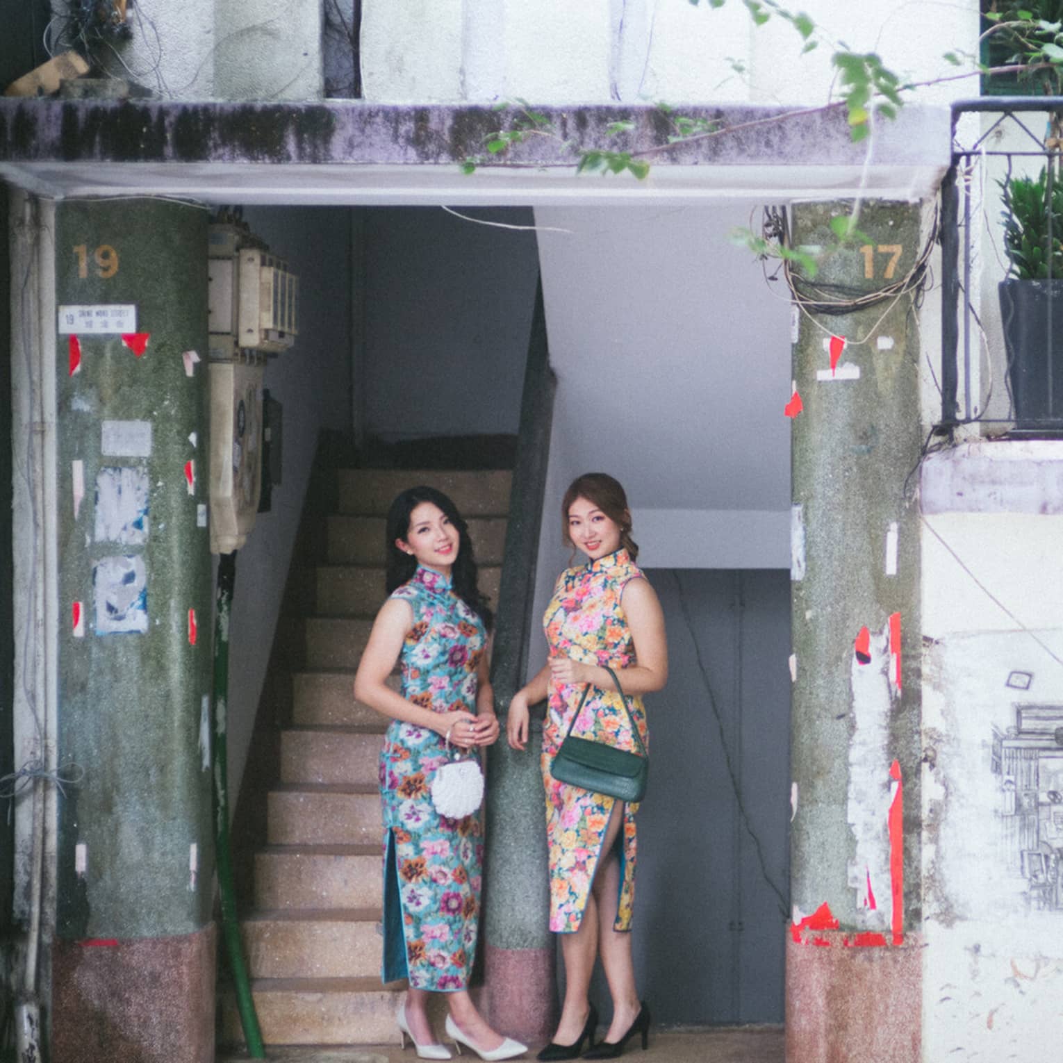 Two people in floral dresses pose on a local unique tour in Central District, holding purses by a staircase with red decorations and wooden pillar.
