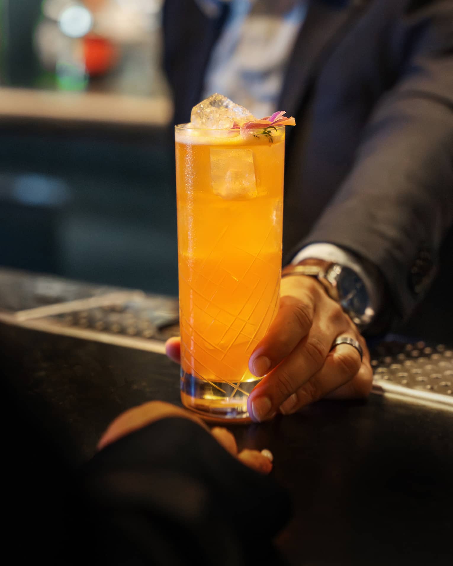 A person sets a bright orange drink in a tall clear glass on a bar counter.