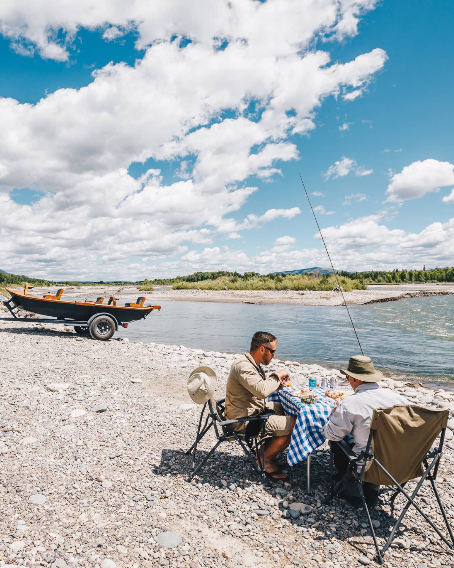 Two fishermen sit at a small table for a lakeside picnic, their boat hitched to their SUV in the background