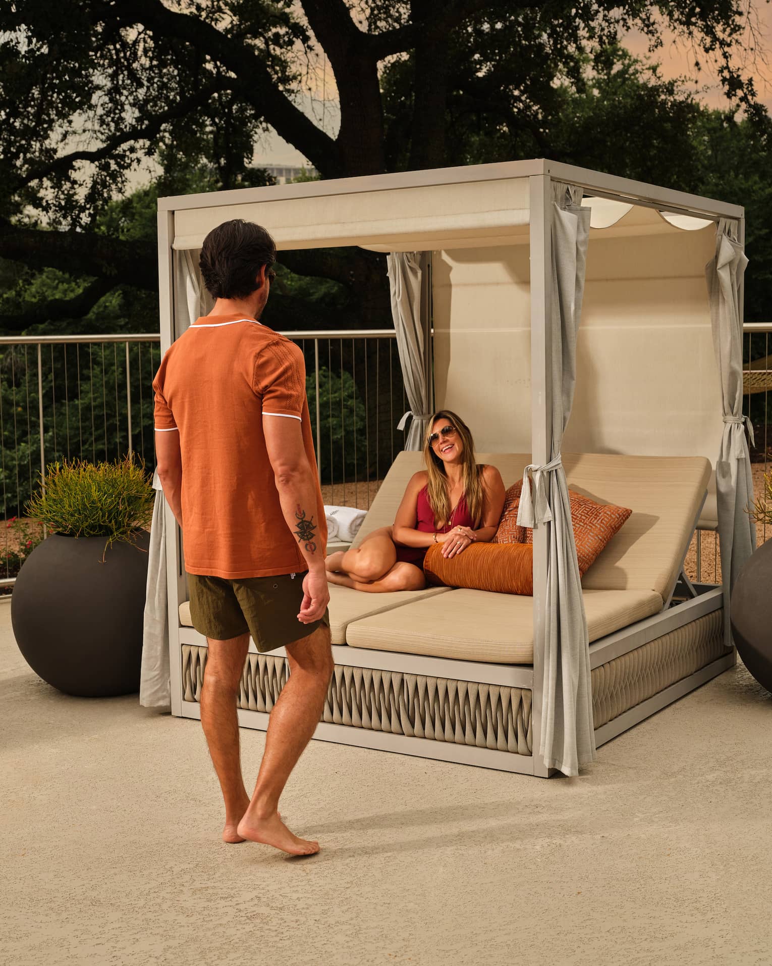 A man walks towards a woman lounging in a cabana surrounded by large potted plants