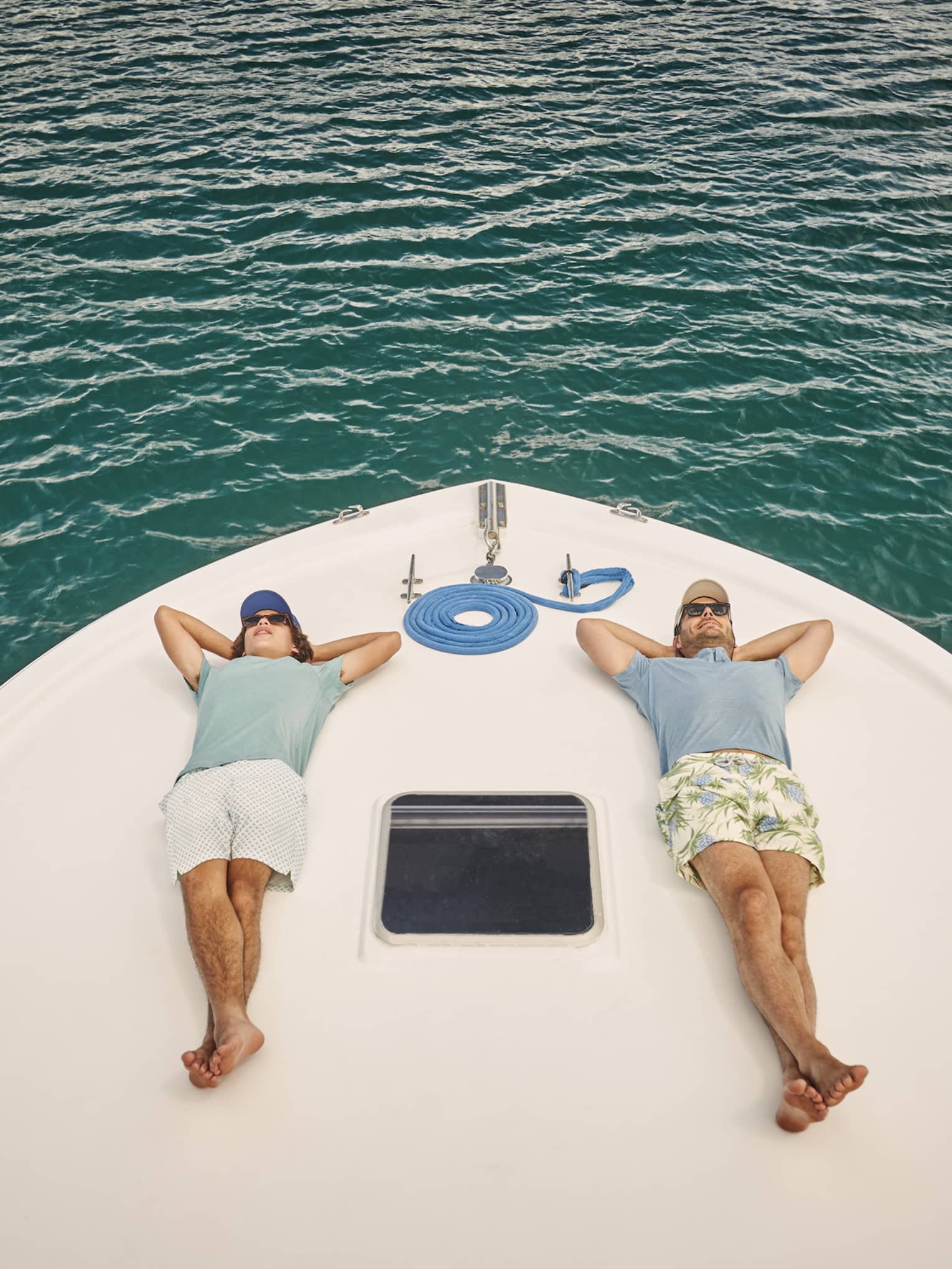 Resting on a boat’s bow over gently rippling water, two passengers lie on their backs, legs crossed, hands behind heads.