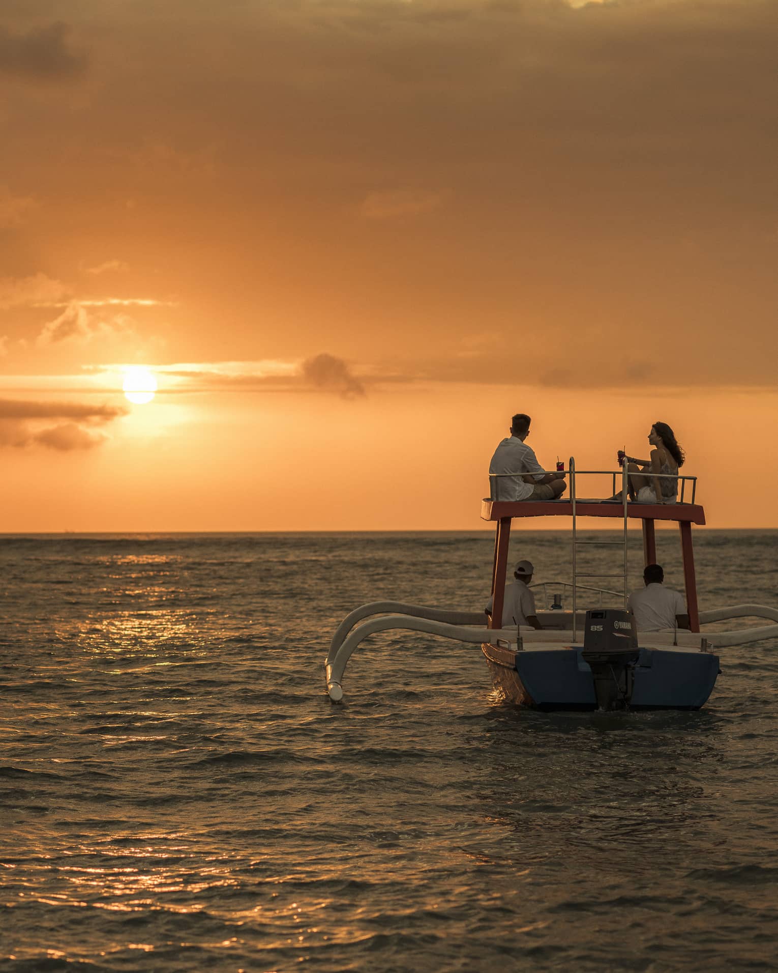 Couple sits on roof of boat sailing on ocean at sunset