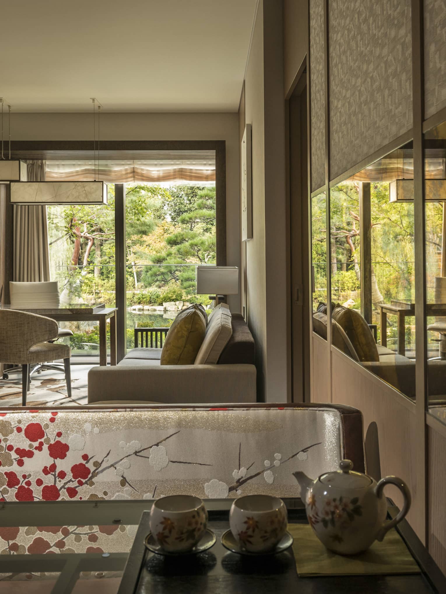 Connecting hotel room with sofa, desk and chairs, floral-patterned screen, tea set and large window overlooking a tranquil pond garden