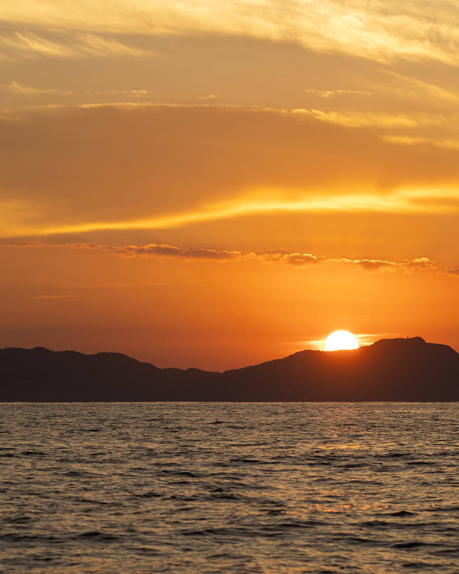 Beyond gently rippling waves, the blazing sun sets behind a hilly island, wispy clouds in the sky awash in its orange glow.