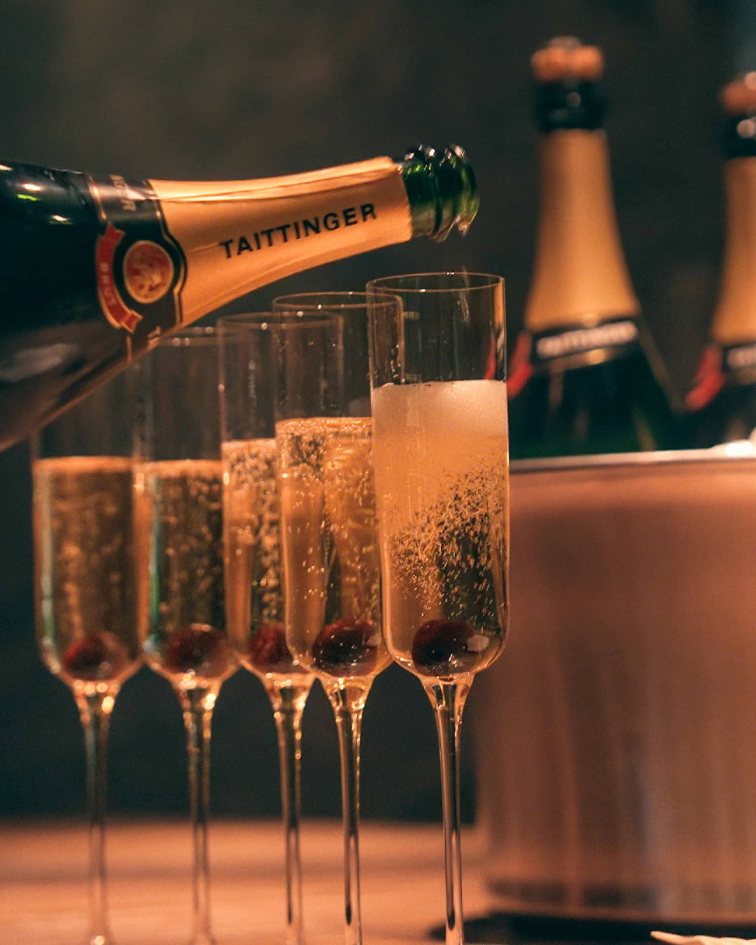 Bottle of champagne being poured into four champagne glasses beside copper ice bucket with three champagne bottles
