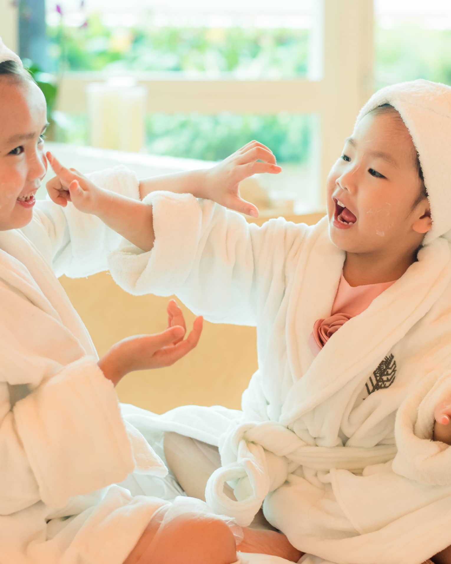 Two laughing young children in white bathrobes with towels around hair put lotion on each other's faces