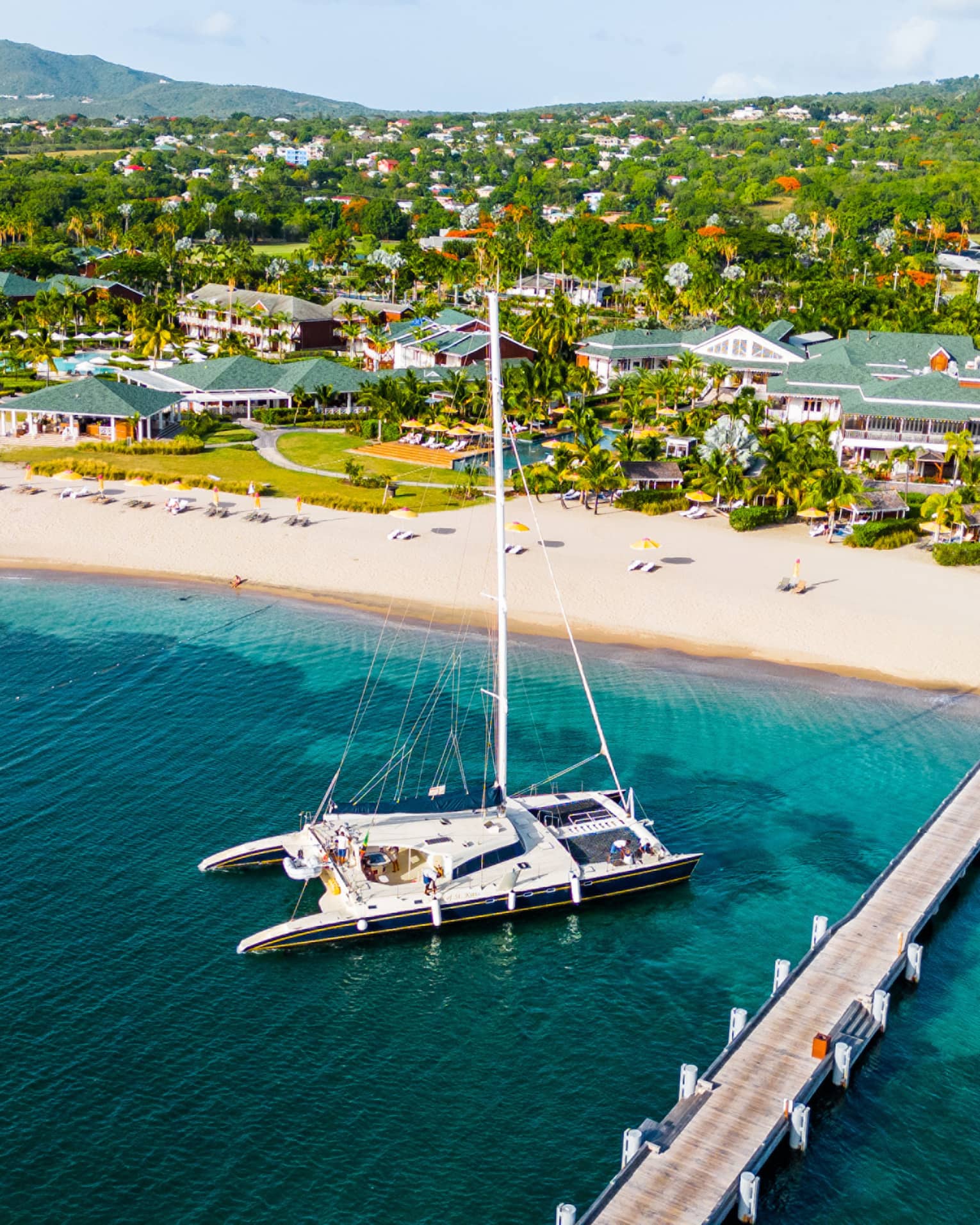 Aerial view of catamaran anchored near a dock reaching out from a beach with a sprawling resort along the coastline set among the trees