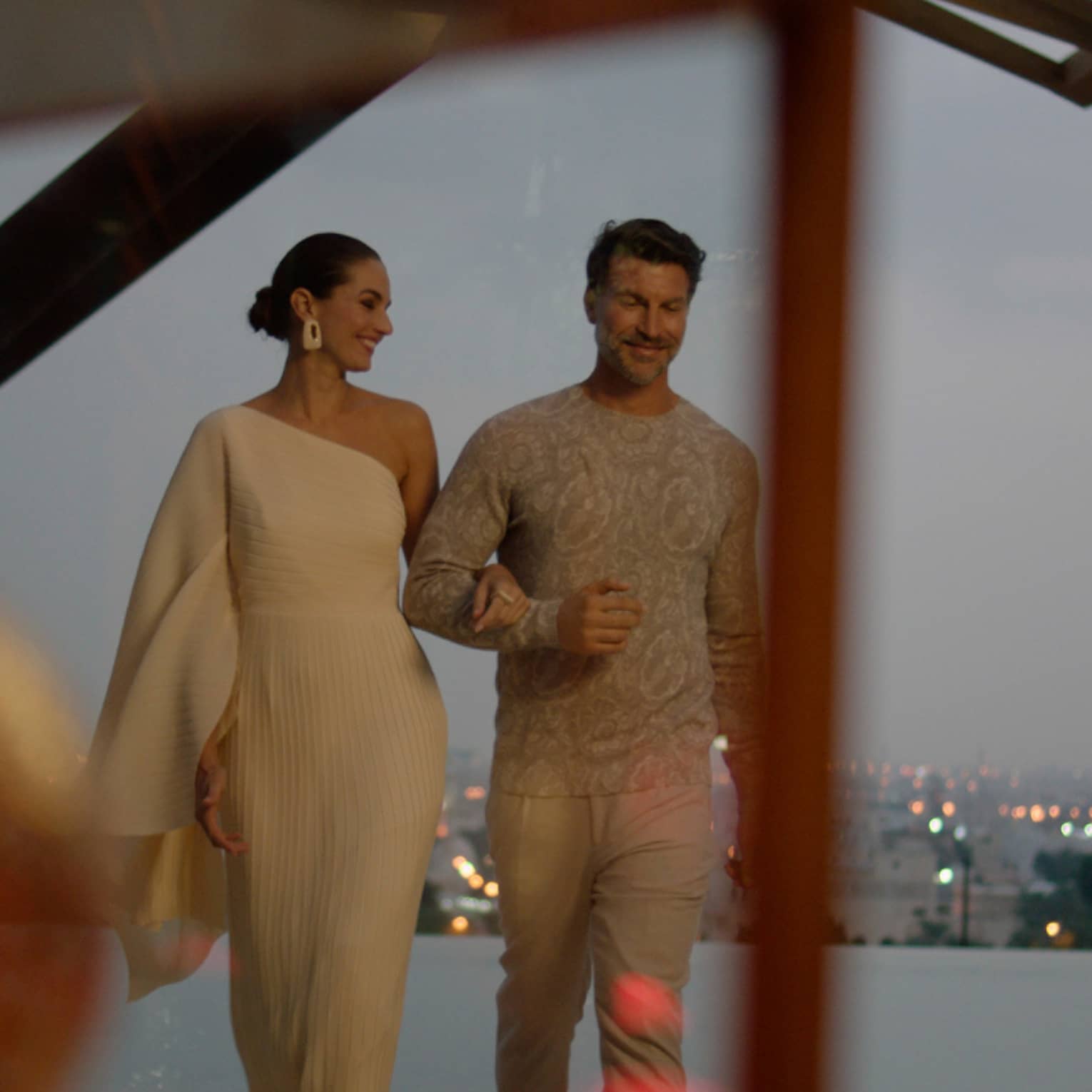 A woman wearing a long white one-shoulder dress walks arm in arm with a man wearing a light-grey sweater and khakis along the edge of a pool with the Kuwait skyline in the background