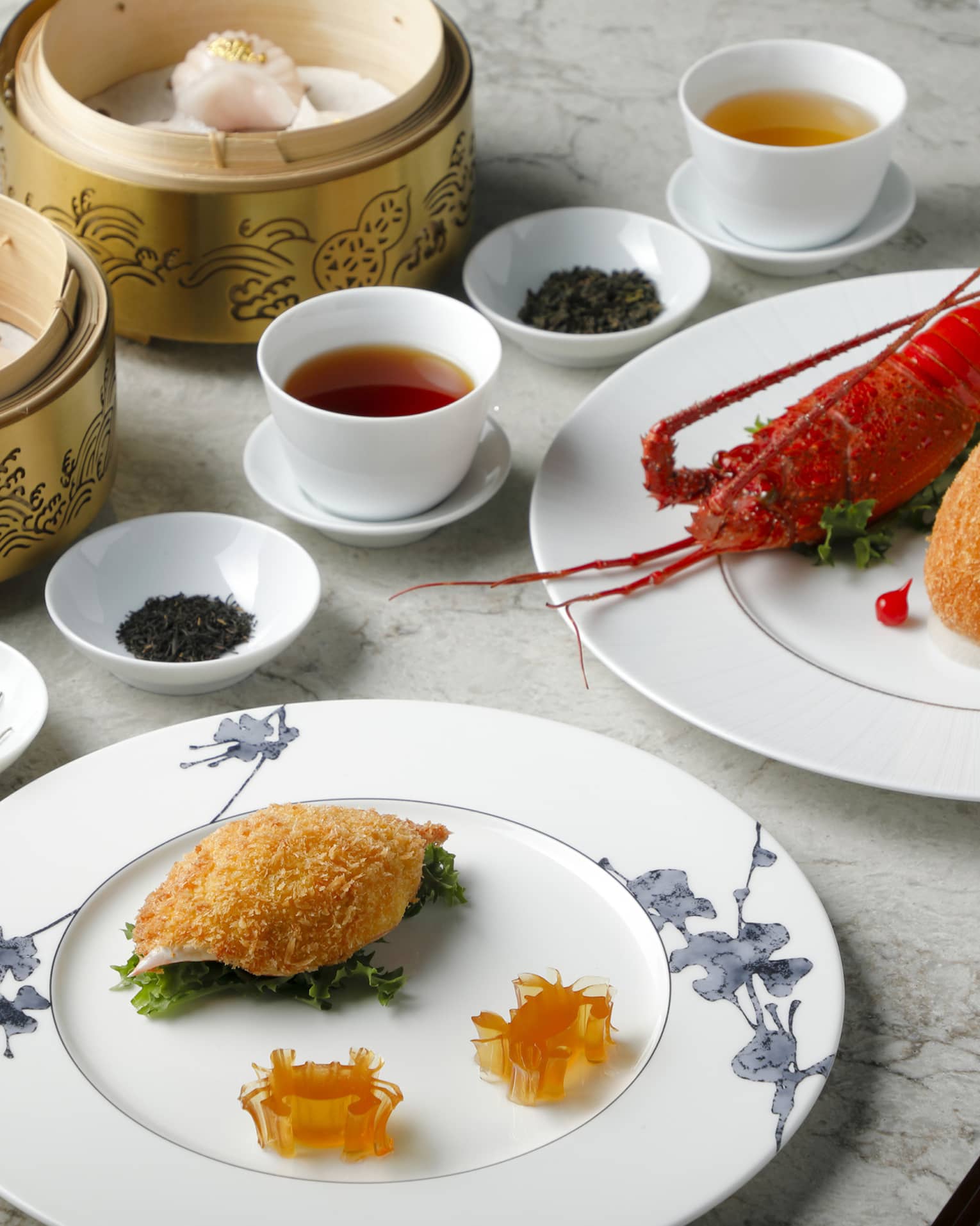 Plain white cups of tea are surrounded by bowls of dried tea leaves, baskets of dumplings and plates of lobster and seafood.