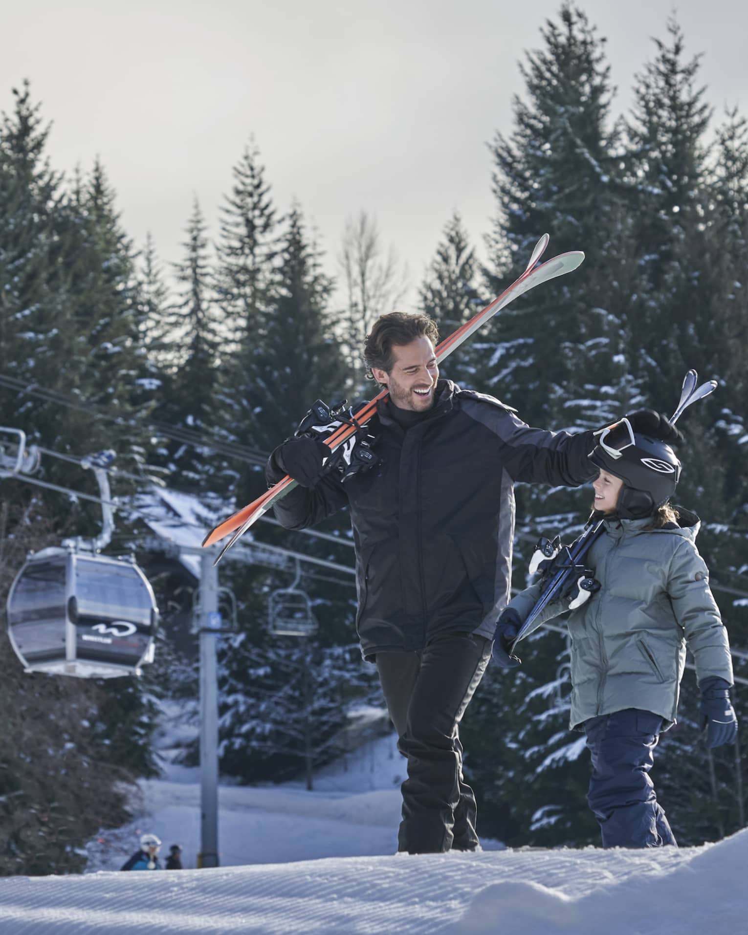 Smiling man and young son carry skis on their shoulders down the slope, ski lift in background 