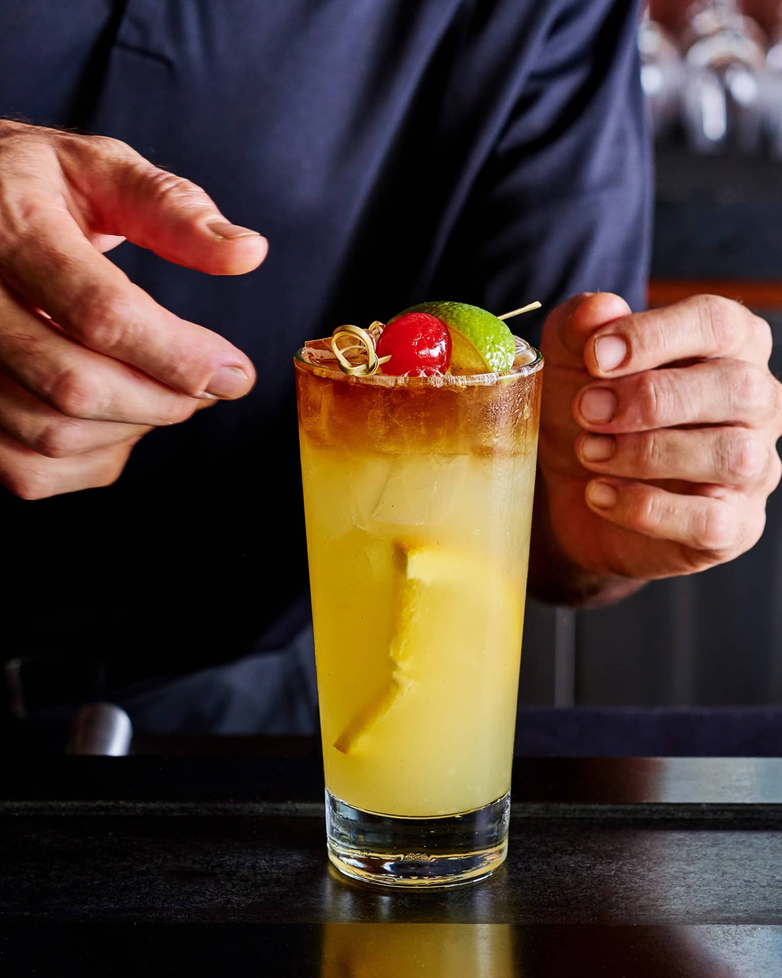 Close-up of hands adjusting a tall orange cocktail on ice, garnished with a cherry and a lime wedge on a small bamboo skewer.