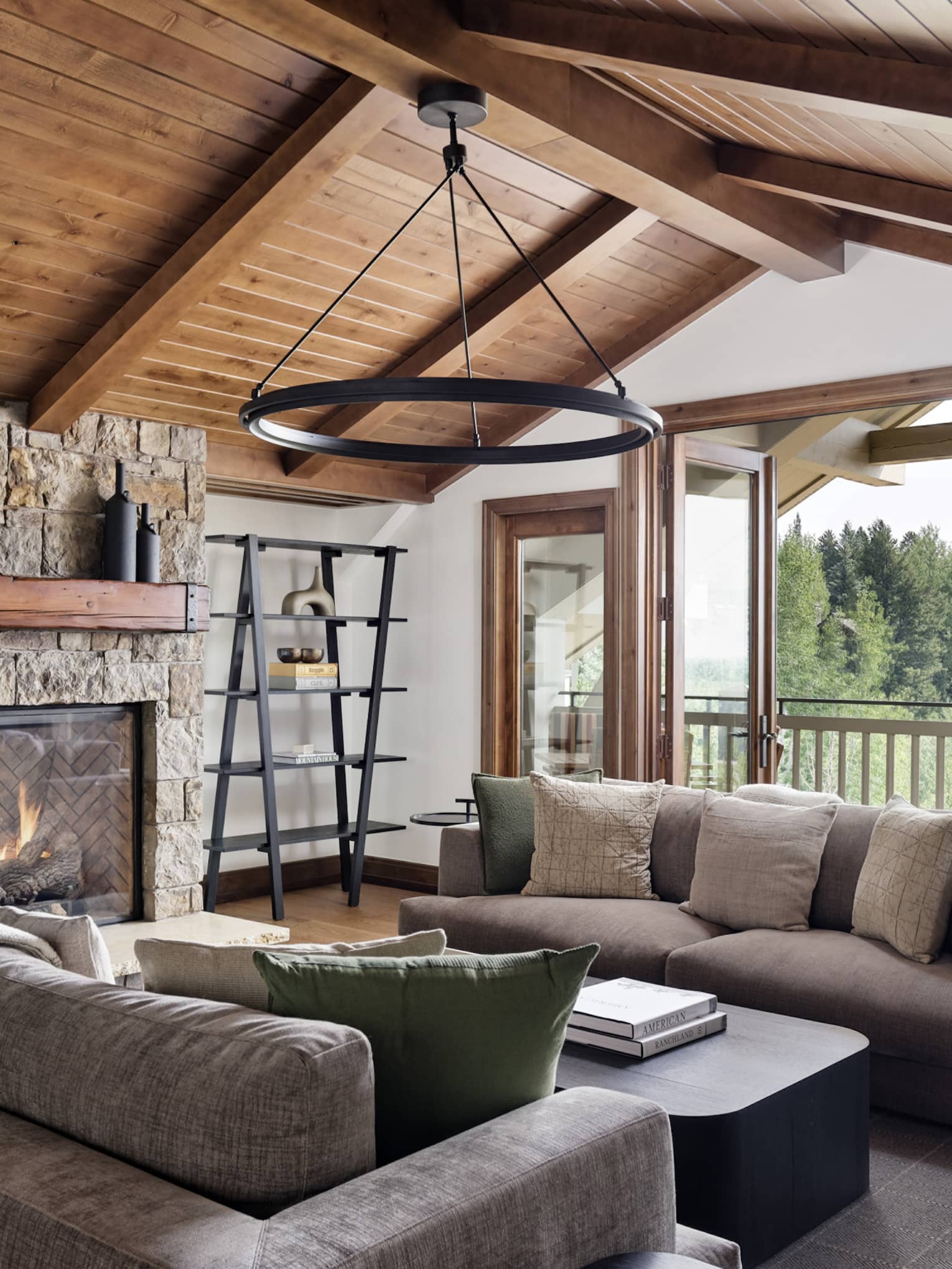 Granite residence living room with vaulted wood ceiling, stone fireplace, grey sofas and a circular chandelier, opening to a balcony with forest view